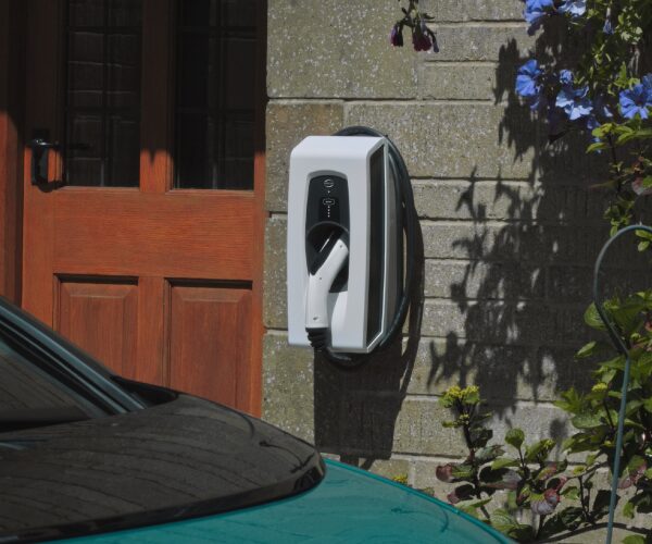 A white Indra wall-mounted EV charger installed on a brick house wall beside a wooden front door, with a car parked in front