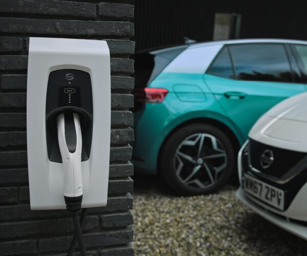 A white Indra electric vehicle wall charger with tethered cable mounted on a dark brick wall, with two electric cars parked on a gravel driveway behind it