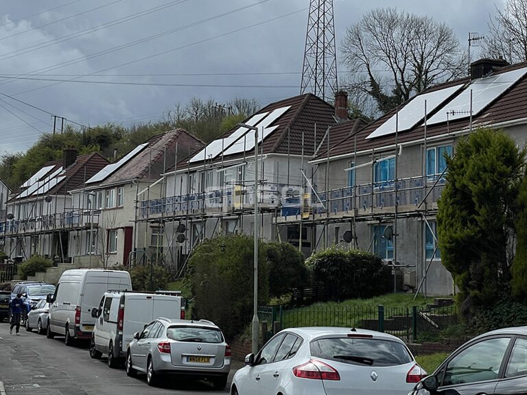 A row of terraced houses with scaffolding and rooftop solar panels being installed, viewed from a residential street with parked cars