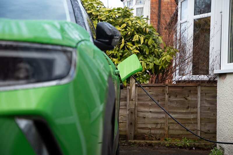A green electric car charging on a UK driveway with a cable plugged into its charging port outside a house
