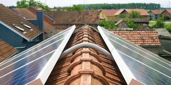 Solar panels mounted on either side of a terracotta tiled roof ridge, with residential rooftops and greenery in the background