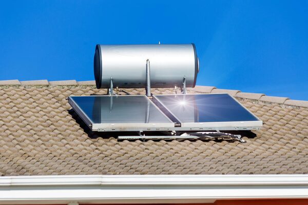 A rooftop solar thermal water heating system with two flat-plate collector panels and a cylindrical hot water storage tank mounted on a tiled roof against a blue sky