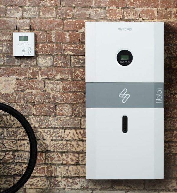 A white myenergi libbi home battery storage unit mounted on a brick wall next to a small libbi controller, with a bicycle wheel in the foreground