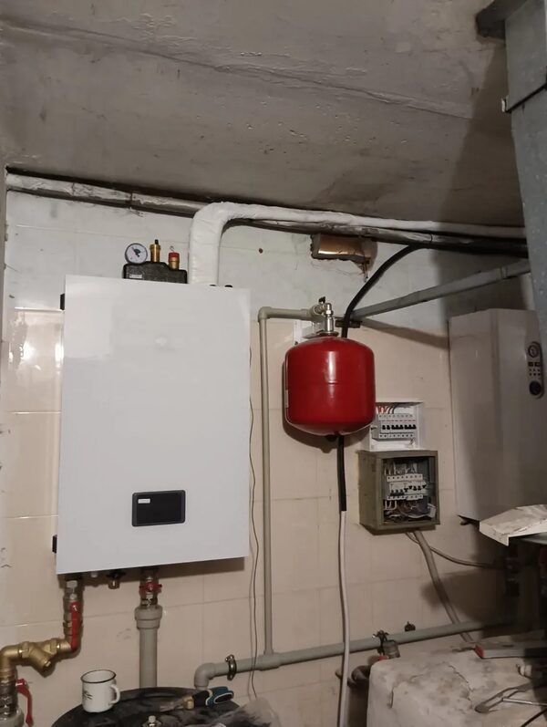 A white wall-mounted heating boiler with a red expansion vessel and pipework installed in a cluttered utility room