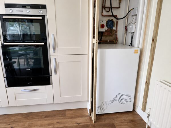A white tepeo ZEB heat battery installed in an unfinished kitchen cupboard next to an oven and exposed pipework