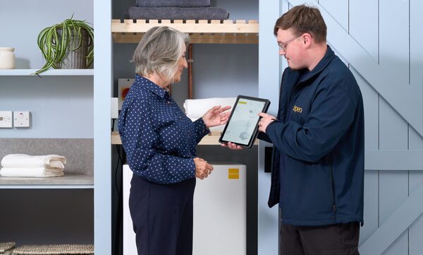 A tepeo engineer in a branded jacket showing a tablet to a woman next to a white tepeo electric thermal battery installed in a utility cupboard