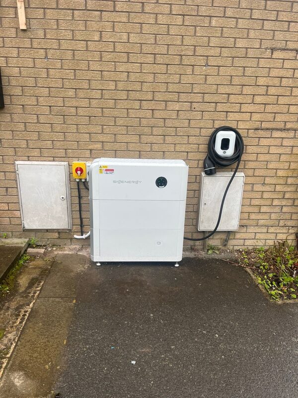 A white Sigenergy home battery unit installed outside against a brick wall next to a wall-mounted EV charger