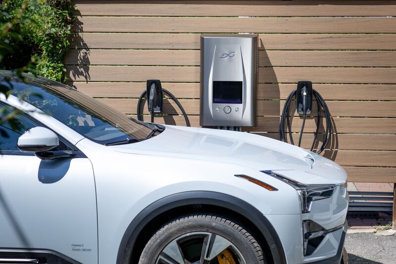 A dcbel Ara bidirectional EV charger mounted on a wooden wall beside a white Polestar 2 electric car