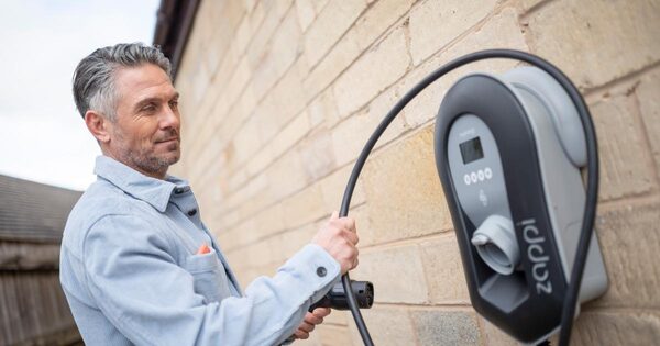A man plugging a charging cable into a wall-mounted Zaptech home EV charge point on a brick house wall