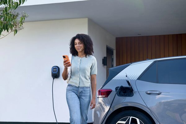 A woman checks her phone beside an electric car charging from a Wallbox wall charger outside a modern house