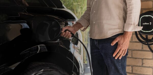A man plugging a charging cable into a black electric car next to a Wallbox EV charger on a brick wall