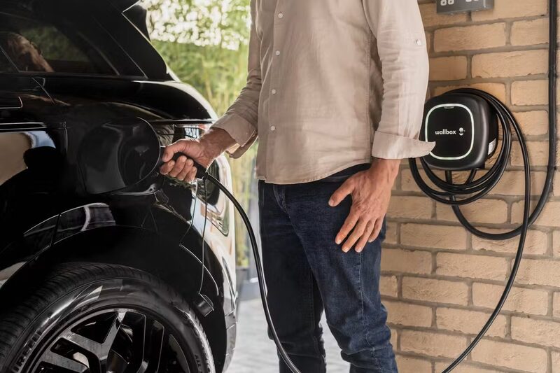 A man plugging a charging cable into a black electric car next to a Wallbox home EV charger mounted on a brick wall