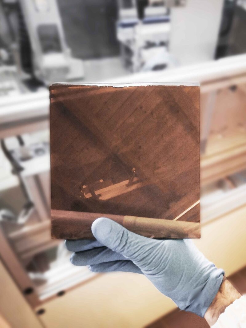 A gloved hand holding a small brown perovskite solar cell sample in a laboratory