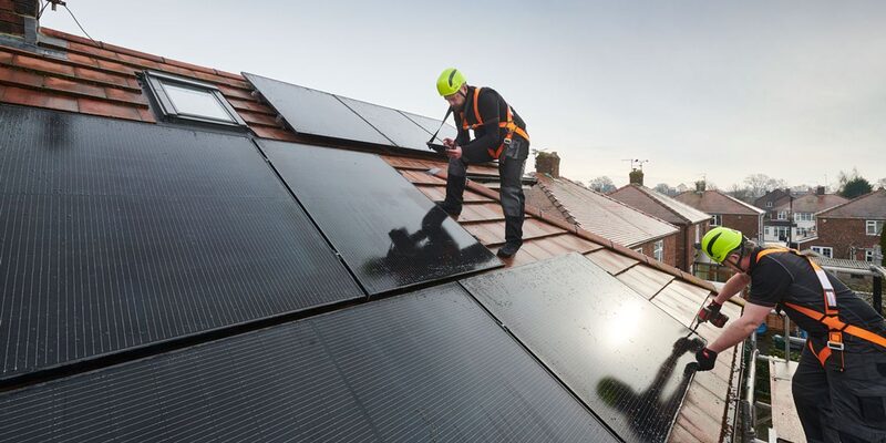Two installers in safety harnesses fitting black solar panels on the tiled roof of a UK house