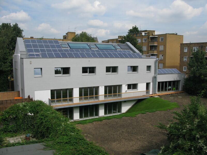 A modern white community centre building with a large rooftop solar panel array and rooflights, surrounded by trees and housing blocks