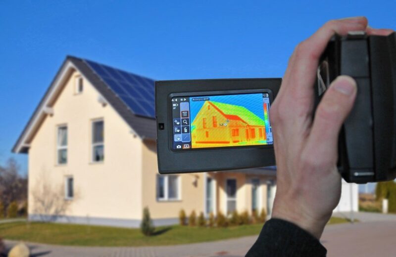 A hand holding a thermal imaging camera photographing a house with solar panels, its screen showing a heat-map of the building