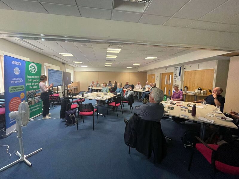 A community energy talk in a village hall, with a presenter speaking to seated attendees beside a Low Carbon Hub banner