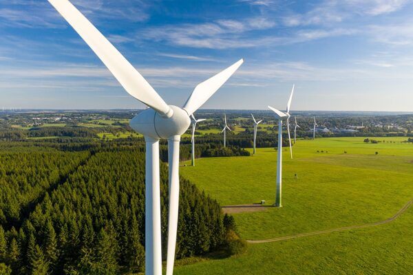 Aerial view of white onshore wind turbines standing in green fields beside a forest under a blue sky