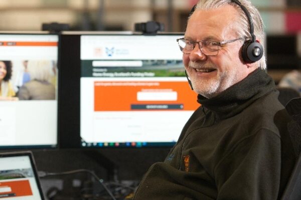 A smiling male Home Energy Scotland advisor wearing a telephone headset sits at a desk in front of computer monitors