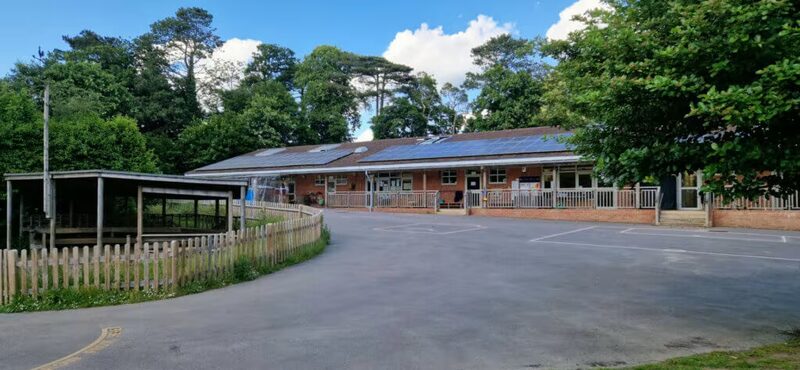 A school building with rooftop solar panels behind a car park and wooden fence