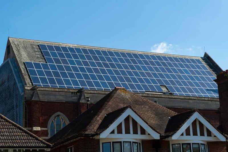 A large array of solar panels installed on the pitched roof of a brick community building, seen above neighbouring tiled house roofs under a blue sky