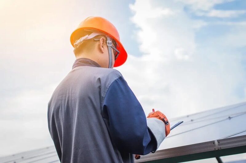 A worker in an orange hard hat inspecting solar panels on a roof while holding a tablet