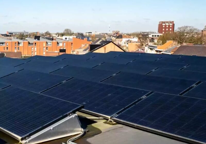 A large rooftop array of solar panels on a flat roof with town buildings in the background