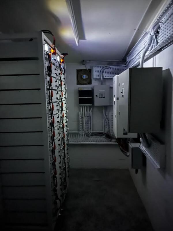 A dark plant room with a large rack of battery storage modules and wall-mounted electrical switchgear and distribution boards