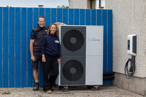 Two people standing beside an outdoor air source heat pump unit next to a house wall with an EV charge point