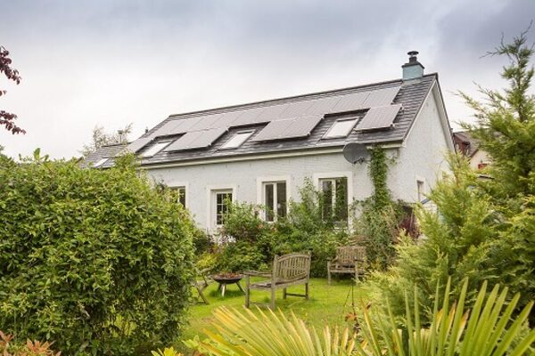 A white rendered house with solar panels on its roof, surrounded by a leafy garden with benches