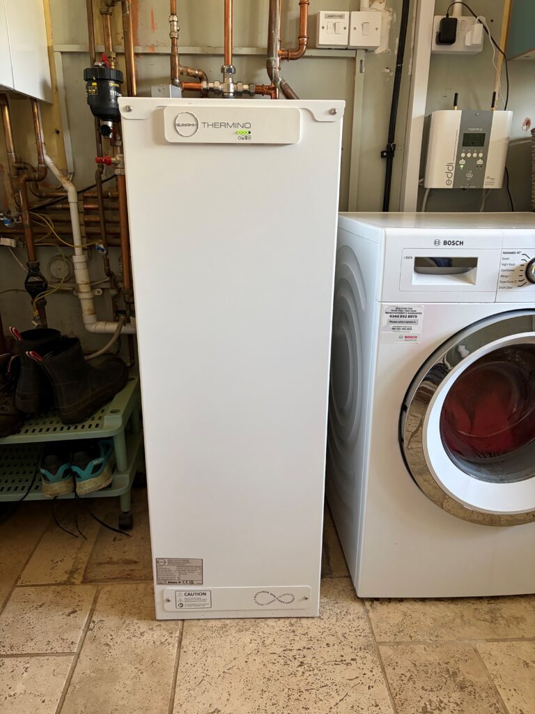 A white Sunamp Thermino heat battery installed in a utility room next to a Bosch washing machine, with copper pipework behind
