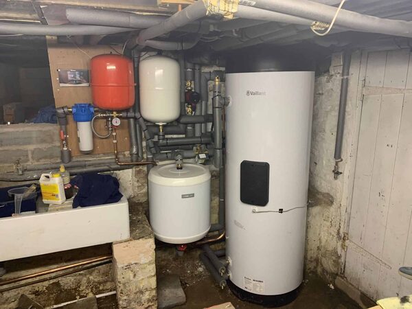 A Vaillant hot water cylinder and buffer tank with expansion vessels and pipework installed in a cluttered cellar utility space