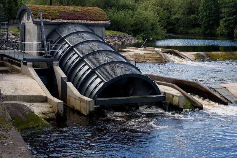 A large hydroelectric Archimedes screw turbine installed at a river weir next to a small turbine house