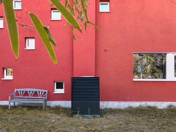 A dark grey outdoor heat pump unit installed against the red exterior wall of a house, next to a wooden bench