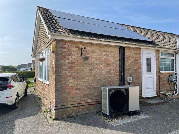 A brick bungalow with rooftop solar panels, an air source heat pump on the outside wall and an electric car parked in the driveway