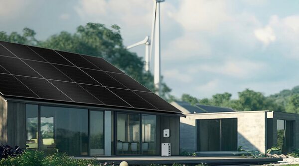 A modern house with black solar panels on the roof, a battery/inverter unit on the wall, and wind turbines in the background