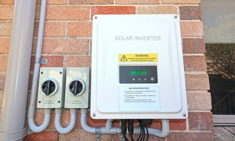 A white solar inverter with warning label and shutdown instructions mounted on a brick wall next to two PV array DC isolator switches
