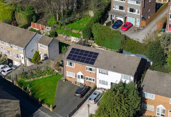 An aerial view of a UK suburban house with solar panels on its roof, surrounded by neighbouring houses and gardens