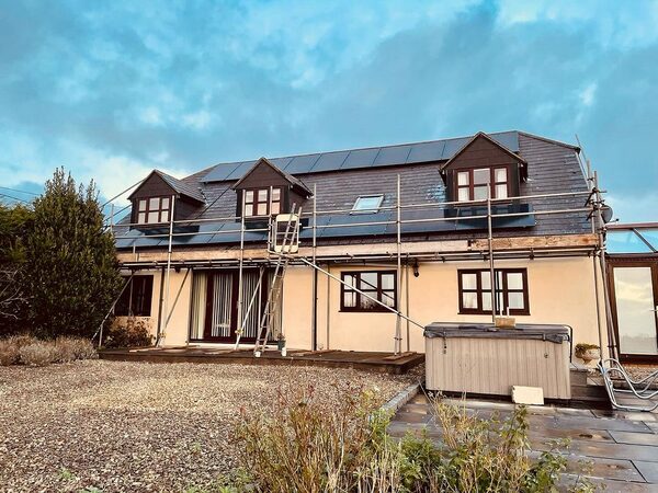 A detached house with solar panels being installed on its roof, surrounded by scaffolding