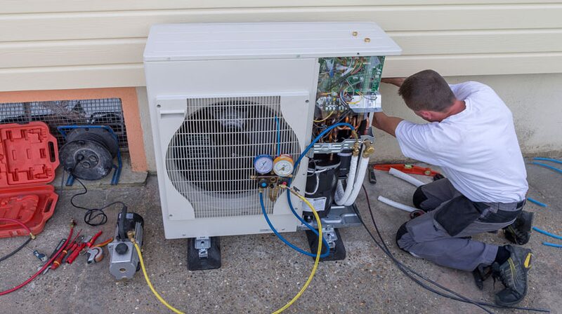 A technician installing and servicing an air source heat pump unit outside a house, with gauges and tools around it