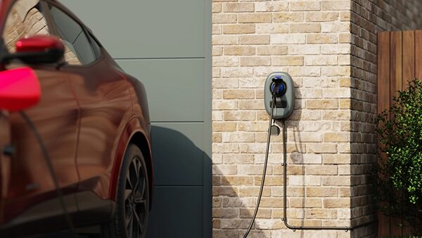 A wall-mounted EV charge point on a brick house exterior with a charging cable running to a parked red car