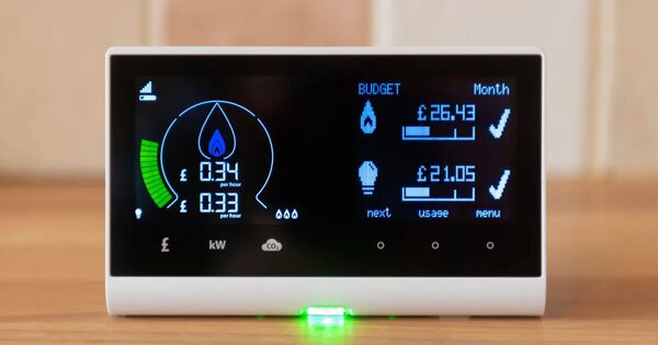 A smart energy monitor in-home display showing live electricity cost per hour and monthly gas and electricity budget usage on a wooden surface