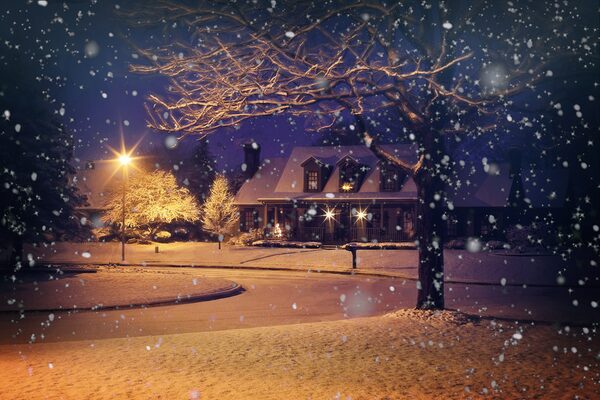 A snow-covered suburban house at night with glowing windows and a street lamp during heavy snowfall