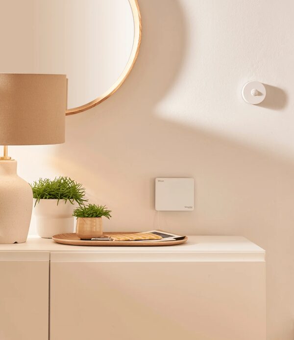 A white Drayton Wiser smart heating controller mounted on a cream wall above a sideboard with a lamp, mirror and potted plants