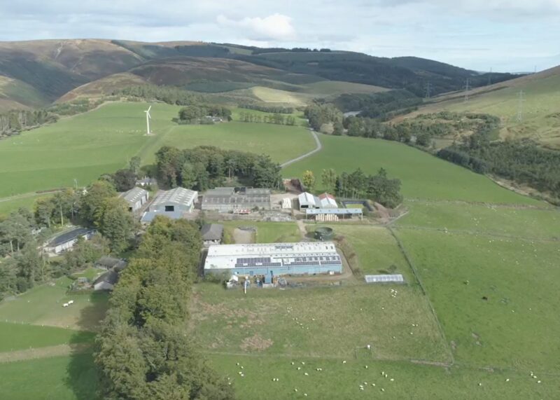 An aerial view of a rural farm with solar panels on a barn roof, a wind turbine and rolling green hills