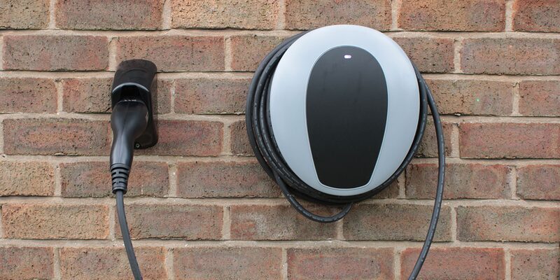 A white and black wall-mounted EV home chargepoint with coiled cable and a holstered charging plug on a brick wall