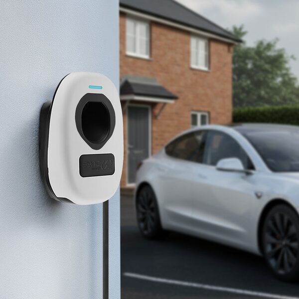A white Sync Energy wall-mounted EV charger on a house wall with an electric car parked outside a brick home