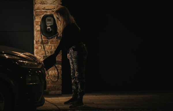 A woman plugging a charging cable into an electric car at night next to a wall-mounted EV charger on a brick wall