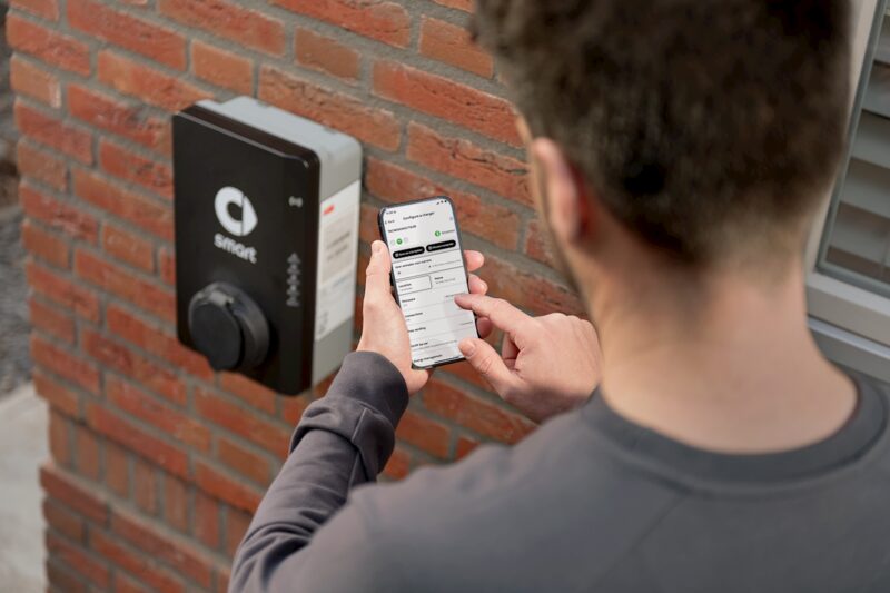 A man using a smartphone app to control an ABB smart wall-mounted EV charger on a brick house wall