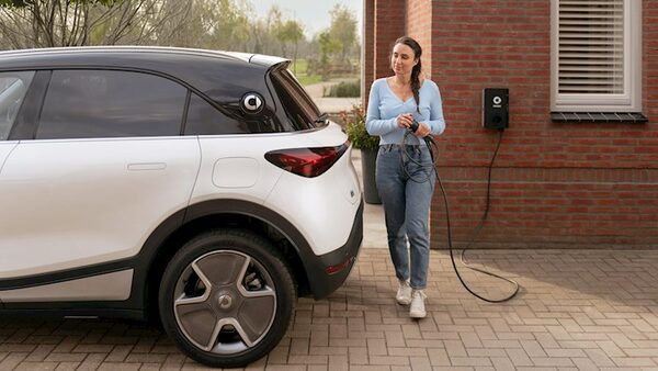 A woman holding a charging cable next to a white electric car plugged into a wall-mounted EV charger on a brick house